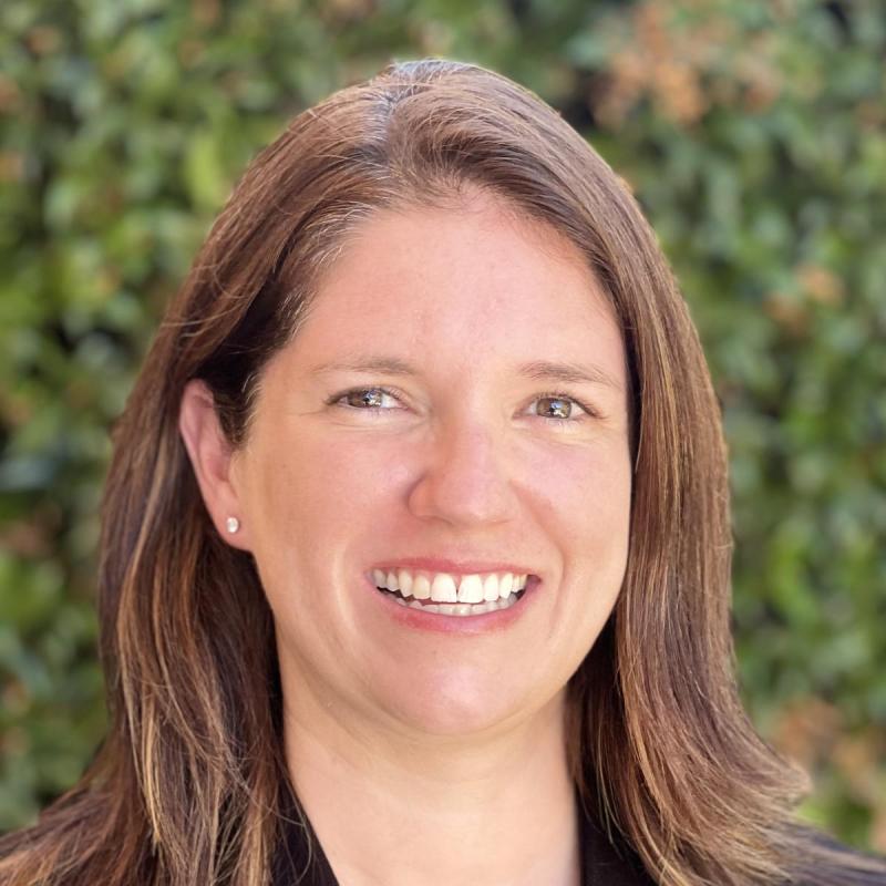 Headshot of Elizabeth Ducy, infront of greenery in a blazer and a blouse with a smile on their face.