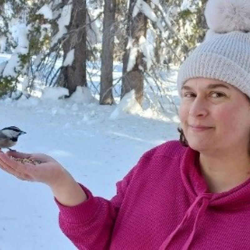 Rachel Donham in a snowy climate wearing a white beanie with a pink hoodie on, smiling, and holding out a palmfull of seeds while an unidentified bird is perched in her hand. 