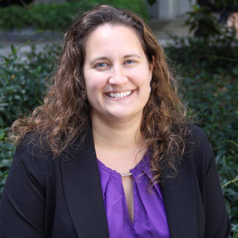 Aja LaDuke sitting infront of greenery wearing a blazer and a purple blouse with her hair slighly pinned back, smiling at the camera. 