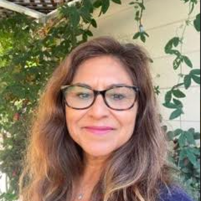 Linda Montes in front of a wall of greenery wearing a blue button up blouse and tortiseshell glasses smiling while wearing her hair down. 