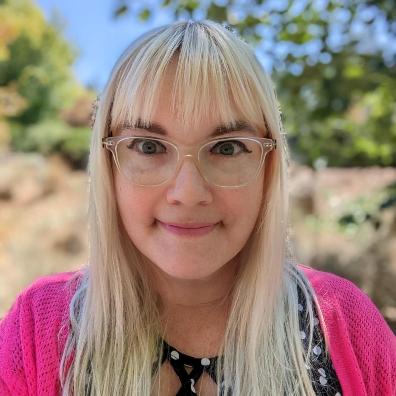 Desirée Poindexter outdoors smiling at the camera wearing a dark blouse with a pink cardigan over top, wearing her blonde hair down with clear framed glasses
