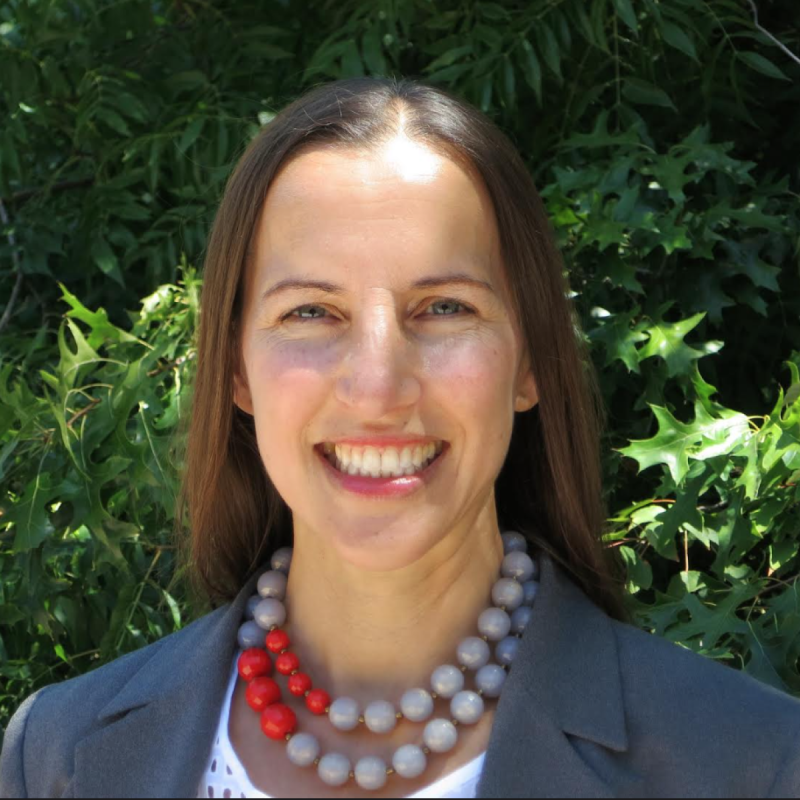 Sarah Rapp standing infront of greenery wearing a white blouse and a dark grey blazer with a grey and red necklace, smiling at the camera. 